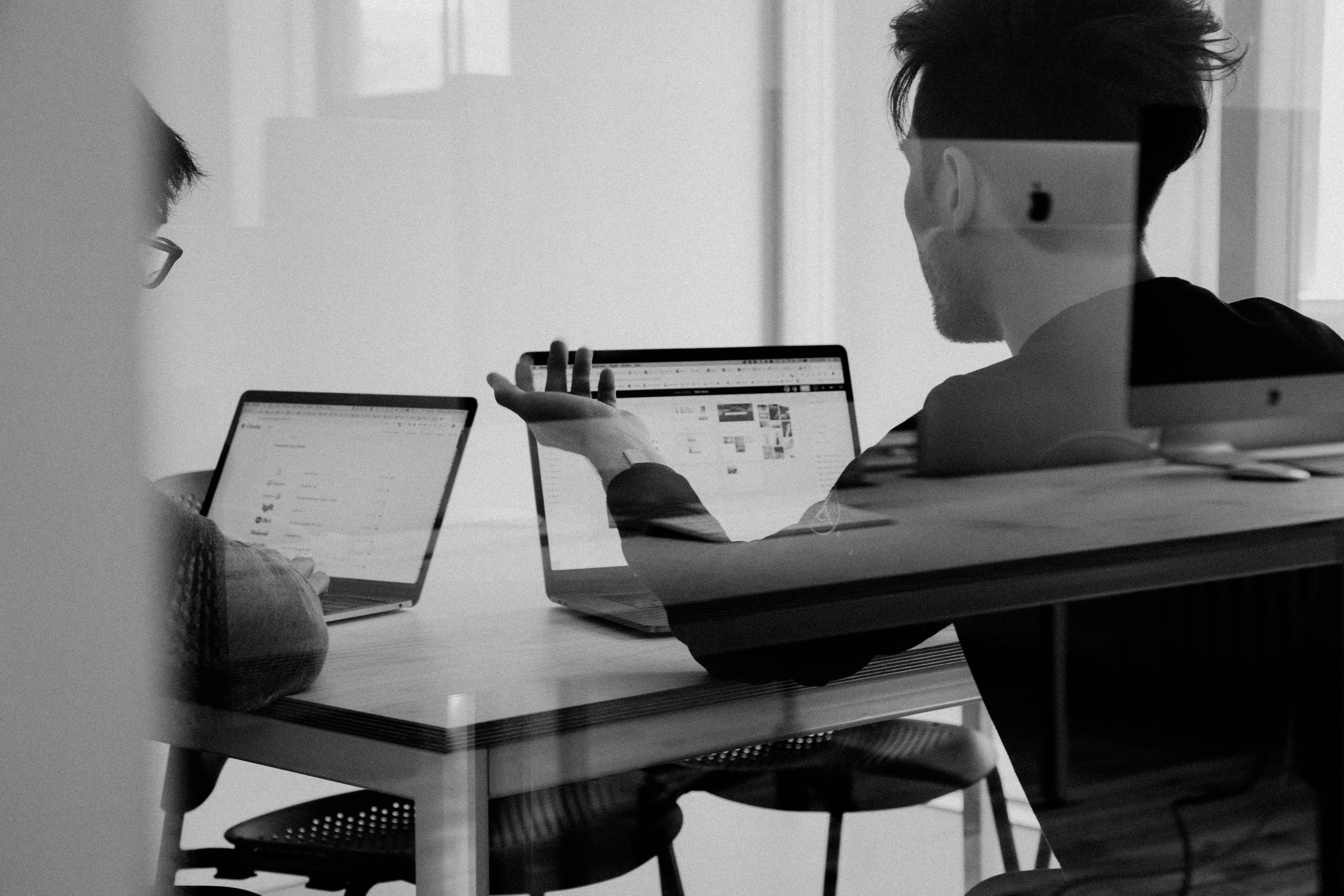 Zwei Personen arbeiten an Laptops an einem Tisch, überlagert von einer leichten Reflexion.