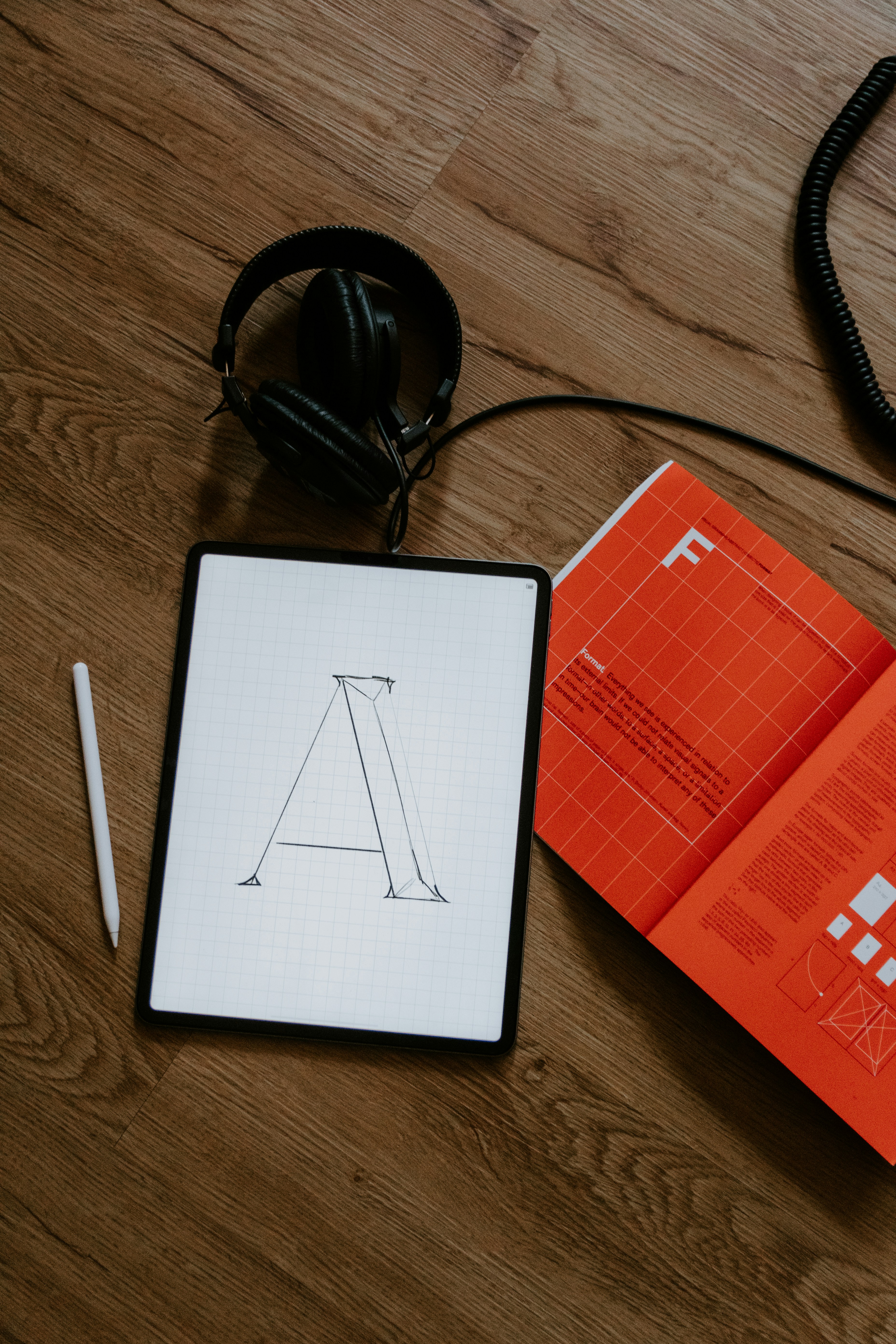 Kopfhörer, ein Tablet mit einem gezeichneten Buchstaben 'A' und ein orangefarbenes Notizbuch liegen auf einem Holzboden.
