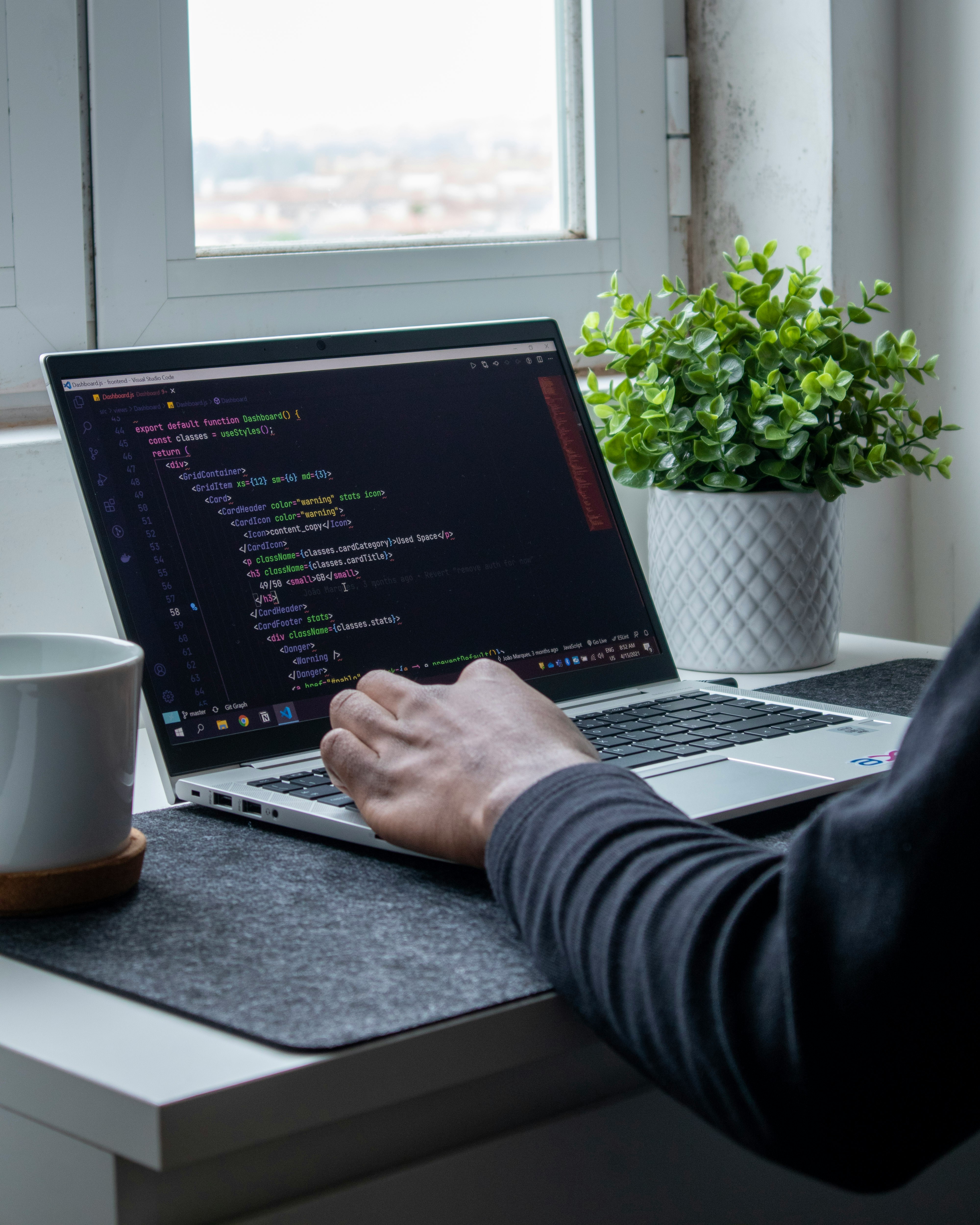 Eine Person tippt auf einem Laptop, auf dem Code zu sehen ist. Daneben stehen eine Pflanze und eine Tasse.