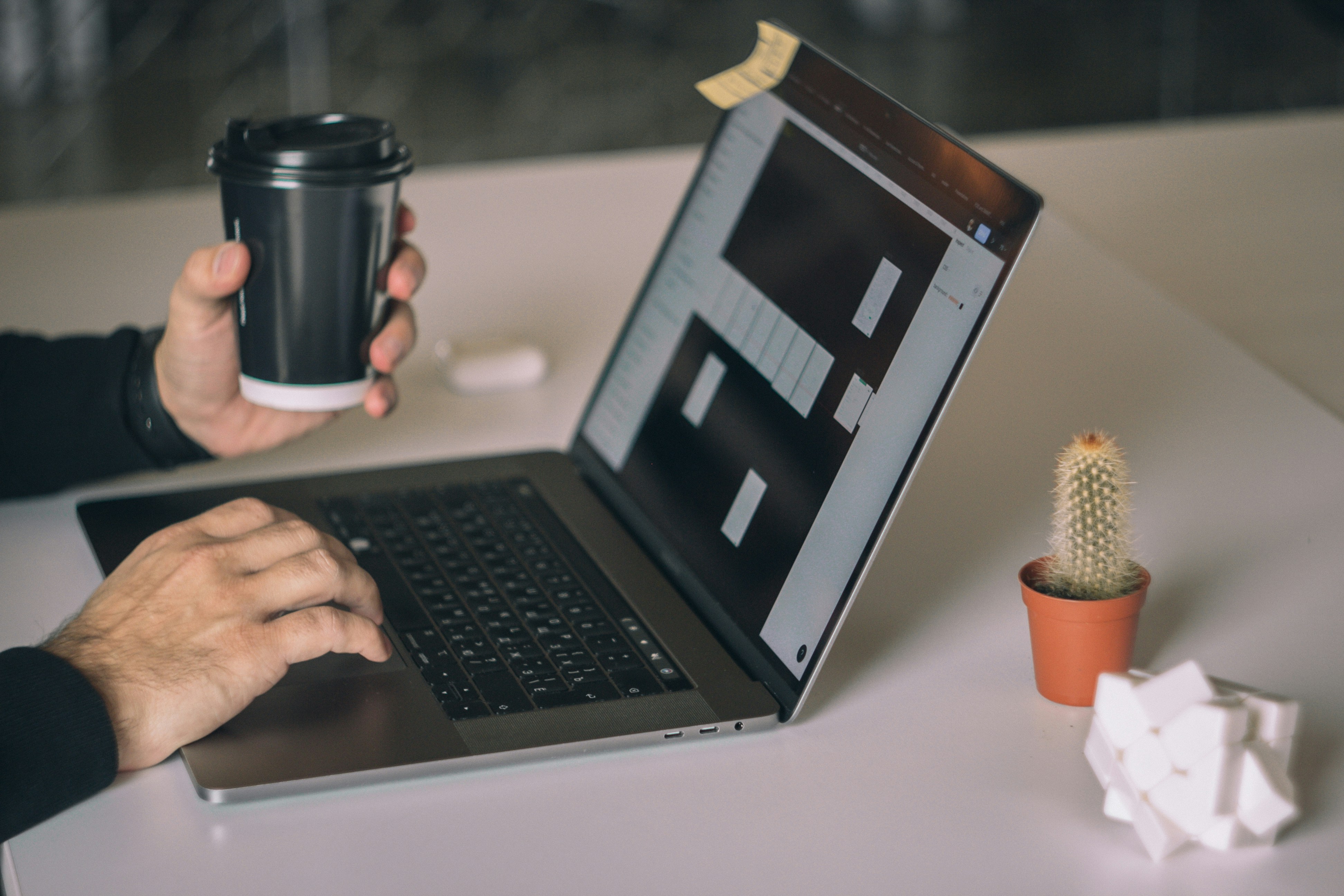 Eine Person tippt auf einem Laptop und hält einen Kaffeebecher. Ein Kaktus und ein Notizzettel sind ebenfalls auf dem Schreibtisch zu sehen.