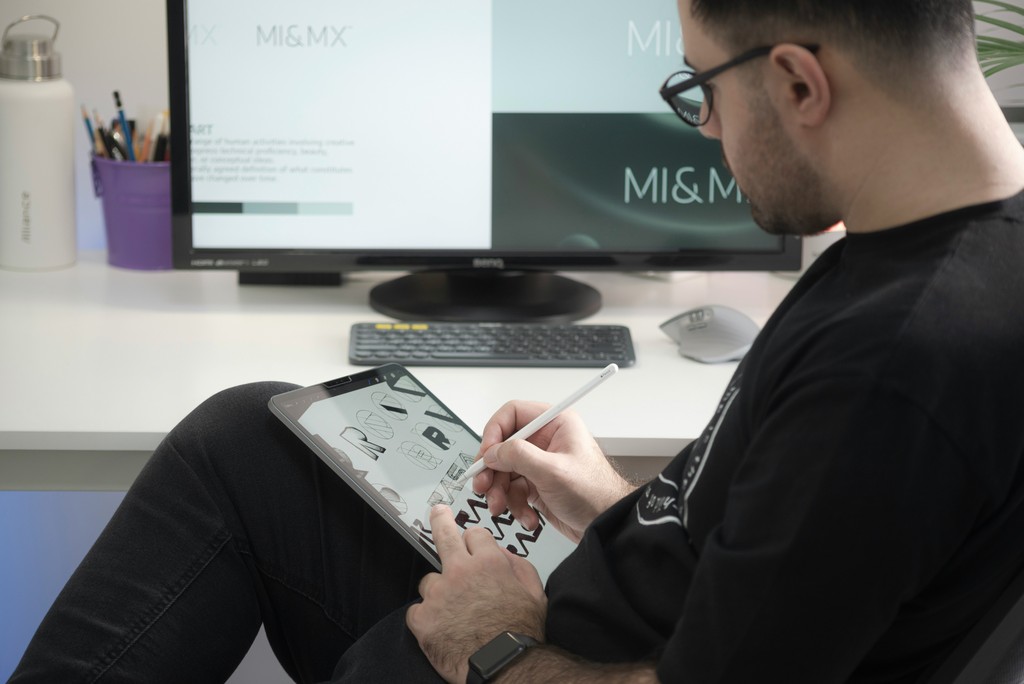 Ein Grafikdesigner zeichnet mit einem Stift auf einem Tablet. Im Hintergrund ist ein Bildschirm mit einem Logo zu sehen.
